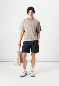 T-shirt en coton rayé beige et marron, shorts bleu marine, associé à un sac fourre-tout beige ; porté avec des chaussettes grises et des baskets.