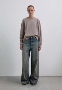 Frau steht in einem taupefarbenen Langarm-Sweatshirt, weit geschnittenen, verblassten blauen Jeans und spitzen braunen Schuhen vor einem schlichten hellen Hintergrund.