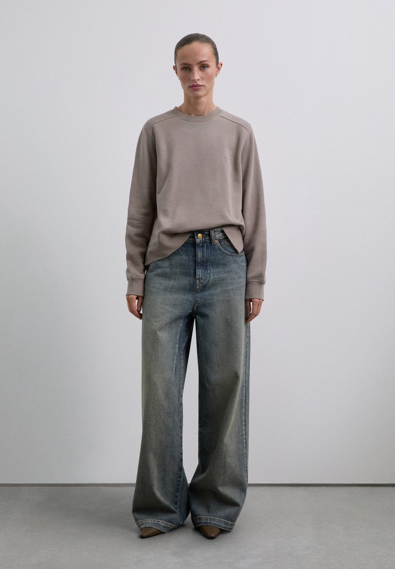 Frau steht in einem taupefarbenen Langarm-Sweatshirt, weit geschnittenen, verblassten blauen Jeans und spitzen braunen Schuhen vor einem schlichten hellen Hintergrund.