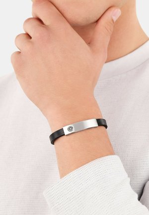 Braccialetto in pelle nera con placca metallica in argento, caratterizzato da un emblema circolare. La texture appare liscia con un design elegante e minimalista.