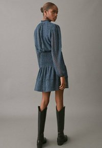 Vestido azul texturizado con mangas largas, cintura fruncida y falda acampanada; combinado con botas negras hasta la rodilla. Vista por detrás.