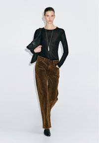 Zwart langemouwen top, bruine corduroy broek en zwarte enkellaarsjes; model houdt een zwarte jas vast. Eenvoudig ontwerp met een strakke, moderne silhouet.