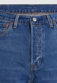 Taille et devant d'un jean en denim bleu avec bouton, passants pour ceinture, surpiqûres orange et poches avant visibles avec rivets.