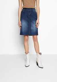 Kvinna i blå denimkjol som går till knäna, beige topp och vita cowboystövlar med klack, stående mot en vit bakgrund.