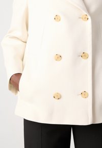 Crème dubbelzijdige blazer met grote ronde schildpadknopen, voorzien van een sjawlkraag en zijzakken, gecombineerd met zwarte broek.