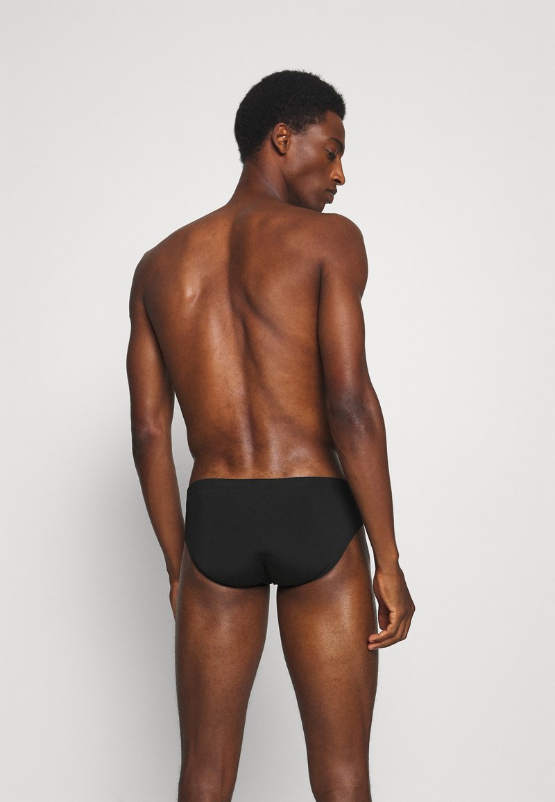 Schwarze Schwimmslips mit glatter Textur und elastischem Bund, getragen von einer Person, die sich abwendet, vor einem neutralen Hintergrund.