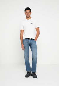 Hvit bomulls-T-skjorte med et lite grafisk motiv, blå denimjeans og svarte treningssko. Avslappet passform og design, stående på en ensfarget bakgrunn.