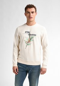 Crème sweatshirt met een botanische graphic, waarop "PTRI Industries" in marineblauw is gedrukt. Zachte textuur en geribbelde manchetten, gecombineerd met blauwe spijkerbroeken.