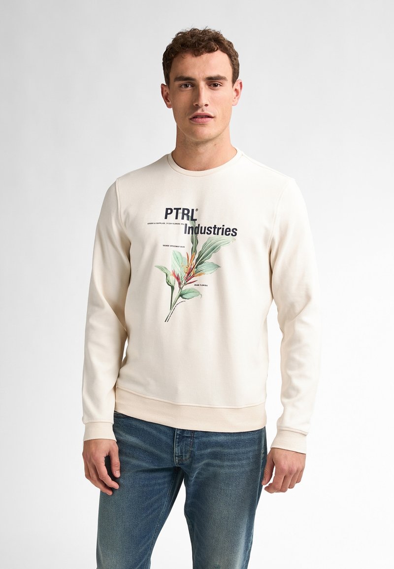 Crème sweatshirt met een botanische graphic, waarop "PTRI Industries" in marineblauw is gedrukt. Zachte textuur en geribbelde manchetten, gecombineerd met blauwe spijkerbroeken.