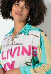 Chemise à boutons colorée présentant un mélange de blanc, turquoise et beige avec un texte en rose et vert vif, des motifs d'étoiles et un design de col.