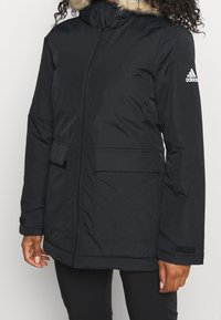 Person in einer schwarzen Adidas Winterjacke mit Kapuze und Fellbesatz, Frontreißverschluss, Klappentaschen und Logo am linken Ärmel.