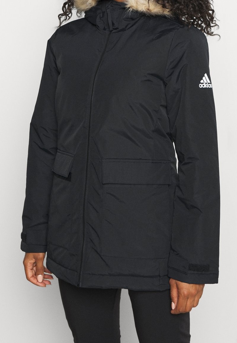 Person in einer schwarzen Adidas Winterjacke mit Kapuze und Fellbesatz, Frontreißverschluss, Klappentaschen und Logo am linken Ärmel.