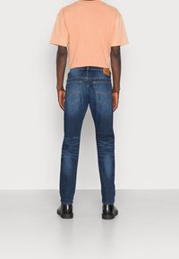 Tmavě modré džínové kalhoty s úzkým střihem, s výrazným designem zadních kapes a koženou hnědou nášivkou s logem, kombinované s broskvovým tričkem.