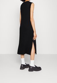 Robe en maille côtelée noire de longueur midi, sans manches, avec une fente latérale, portée avec des chaussures noires épaisses et des chaussettes blanches.