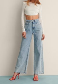 Hellblaue, weit geschnittene Jeans mit hoher Taille, aus Denim-Material und kontrastierenden Nähten, kombiniert mit einem weißen Cropped-Top und roten Riemchen-Pumps.