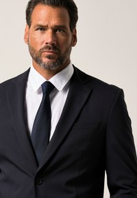 Giacca da uomo blu scuro con revers a punta, abbinata a una camicia bianca e a una cravatta scura a trama, che mette in risalto il design classico dell'abbigliamento maschile.