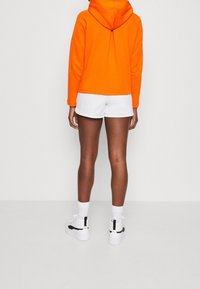 Persoon in feloranje hoodie, witte shorts, witte sokken en witte sneakers met zwarte accenten, staand met zijn rug naar de camera op een effen achtergrond.