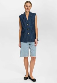 Tumesinise triibulise ilma varrukateta bleiseri kuldsete nööpidega, koos heledate teksashortsidega, millel on kulunud äär, kantud mustade lamejalanõudega.