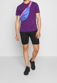 Maglietta viola, pantaloni corti neri e sneakers neri. Una cintura azzurra con dettagli rosa e un logo nero è indossata a tracolla.