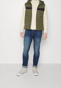 Grüner gepolsterter Weste mit Kapuze, cremefarbener Zopfstrickpullover, blaue Jeans und graue High-Top-Sneaker mit weißen Sohlen.