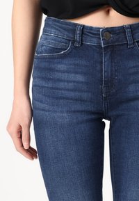 Jeans de mezclilla azul oscuro con un diseño ajustado, que cuentan con bolsillos estándar, cierre de botón y una ligera textura desgastada en la parte delantera.