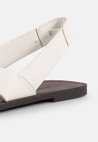 Baltos odinės šlepetės su plačiomis dirželis ir plokščiu rudu padu. Pasižymi minimalistiniu dizainu ir tekstūruota paviršiumi viršuje.