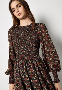 Sukienka z długimi rękawami z czarnego materiału z kwiatowym wzorem w odcieniach czerwieni, żółci i zieleni. Fason z marszczonym biustem, rozkloszowanym dołem oraz marszczonymi rękawami.