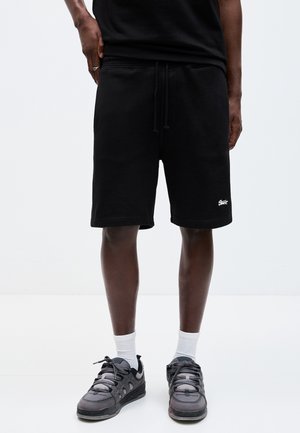 Man wearing black drawstring shorts with white logo near hem, white socks, and black sneakers, standing against plain white background.