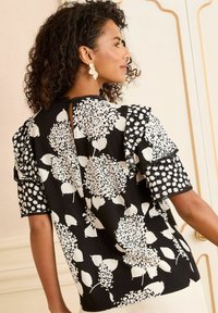 Blusa nera con audaci motivi floreali bianchi, maniche a sbuffo e schiena a chiave. Delicato bordo in pizzo valorizza le maniche.