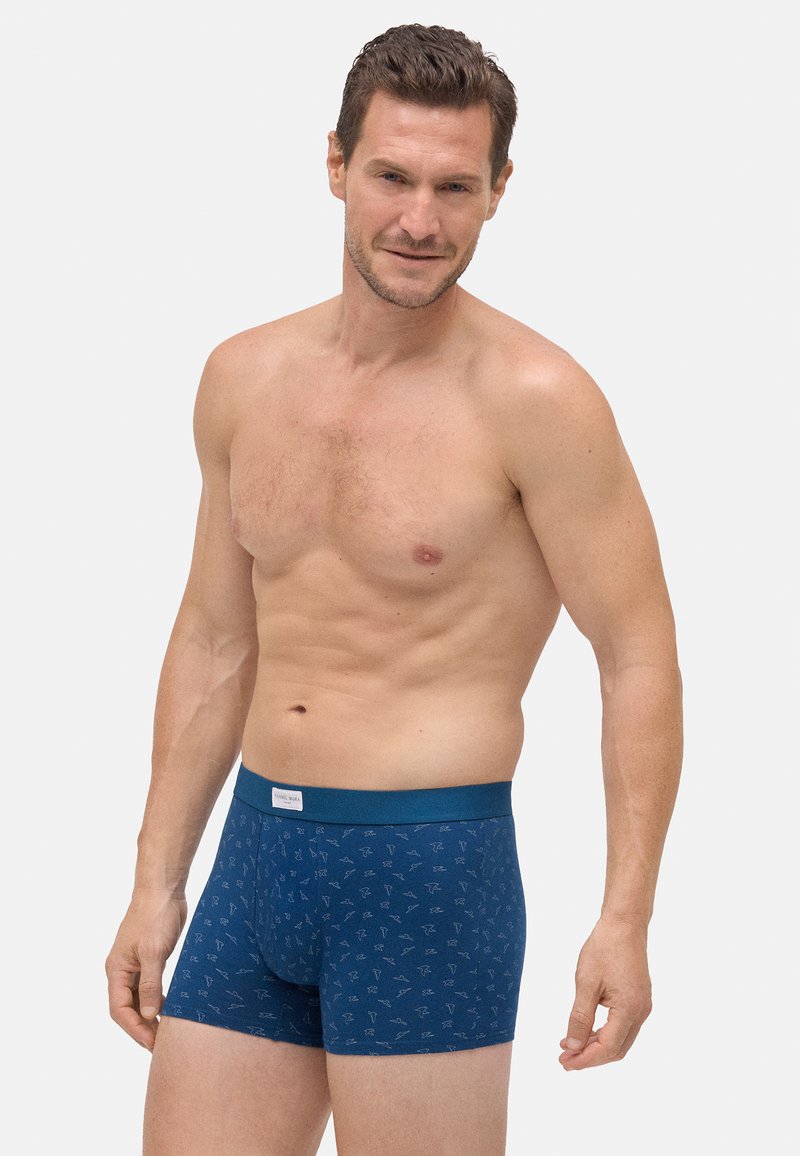 Hombre con cabello corto castaño que lleva calzoncillos de cuadros azules contra un fondo blanco liso, posando con los brazos relajados.