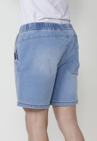 Mees, kes kannab heledat sinist denimist lühikarvadega pükse, millel on elastne vöökoht ning taskud, koos valge T-särgiga, lihtsa tausta ees.