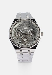 Relógio octagonal prateado com um mostrador texturizado e cintilante, três submostradores, detalhes em cristal e uma bracelete metálica em elos. Marca: Guess.