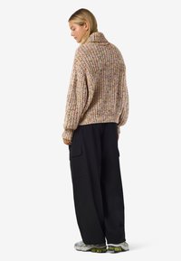 Mulher vestindo um suéter de malha volumosa multicolorido, calças cargo pretas largas e tênis brancos, de pé, virada de lado, em um fundo branco.