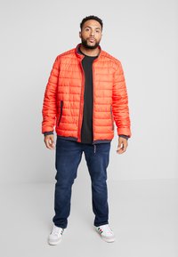 Felle oranje pufferjack met een gewatteerde textuur, een ritssluiting aan de voorkant en marineblauwe accenten. Draagt over een zwart shirt, gecombineerd met blauwe jeans en witte sneakers.