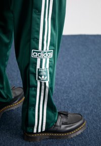 Green athletic pants with white three-stripe design, featuring snap buttons. An Adidas logo patch and a "Brand with Three Stripes" label are visible.