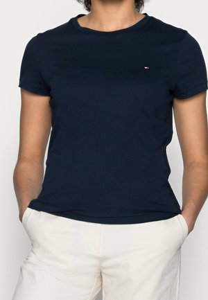 Persona con camisa de manga corta azul marino con un pequeño logo en el pecho y pantalones blancos, con las manos en los bolsillos, fondo neutro.