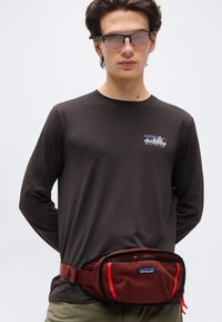 Νεαρός άνδρας που φοράει σπορ γυαλιά ηλίου με χρωματιστούς φακούς, μια μαύρη μπλούζα μακρυμάνικη Patagonia και μια κόκκινη τσάντα μέσης Patagonia σε λευκό φόντο.