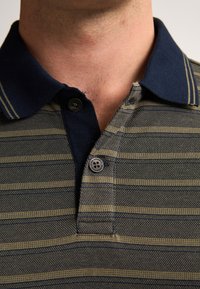 Polo tričko s límečkem s námořnicky modrými akcenty a olivově zeleným a námořnicky modrým pruhovaným vzorem. Dva knoflíky na výstřihu, strukturovaná tkanina.