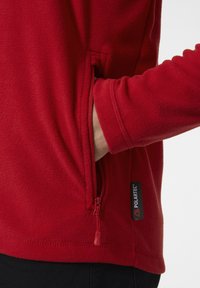 Czerwony polarowy sweter z boczną kieszenią na zamek, logo Polartec oraz miękką teksturą. Design ma dopasowany krój i długie rękawy.