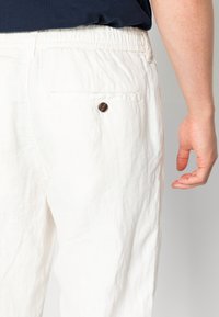 Vita linnebyxor med elastisk midja, som har en bakficka med en brun knappdetalj. Strukturerad, lättviktsmaterial.