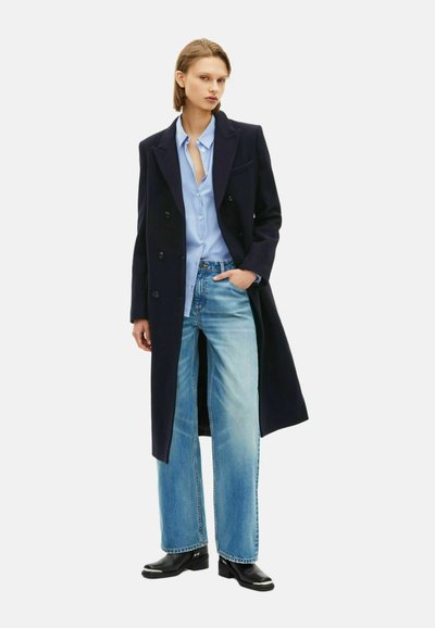 Marineblauwe lange jas over een lichtblauwe blouse, gecombineerd met wijde, vervaagde jeans en zwarte enkellaarsjes. Bevat dubbelzijdige knopen en heeft een relaxte pasvorm.