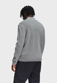 Man met zwart getwist haar, gekleed in een grijze gebreide trui en zwarte broeken, staand met zijn rug naar de camera tegen een effen achtergrond.