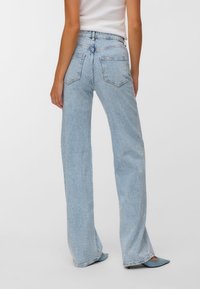 Jeans de mezclilla azul claro de talle alto con ajuste de pierna recta, patrón texturizado sutil y dos bolsillos traseros. Combinados con zapatos azules de punta estrecha.