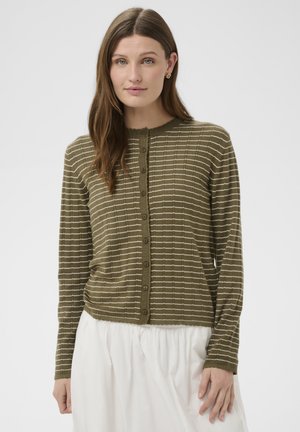 Vrouw die een olijfgroene, gestreepte cardigan met knopen en een witte rok draagt, staande tegen een effen lichtgrijze achtergrond.