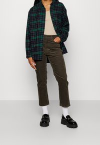 Chemise oversize à carreaux verts et bleu marine, débardeur beige, pantalon en velours côtelé vert olive, chaussettes blanches, mocassins à plateforme noirs avec détail chaîne.