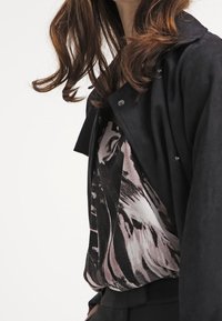Kobieta ubrana w czarną kurtkę na wzorzystej czarno-białej bluzce, z luźnymi brązowymi włosami opadającymi na ramiona.