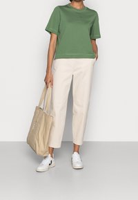 Persona con un top verde a maniche corte, pantaloni cropped beige, sneakers bianchi, che tiene una borsa a tracolla beige con testo sulla tasca, in piedi su un pavimento grigio.