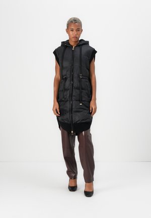 Black sleeveless puffer vest with a hood, front zipper, side pockets, and drawstring waist, paired with dark brown tapered pants and heels.