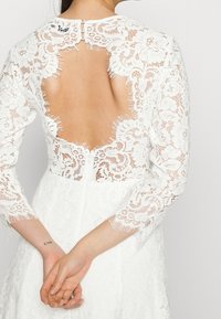 Valkoinen pitsimekko, jossa on pitkät hihat ja syvä selkäaukko. Suunnittelussa on kukkakuvioita ja revittyjä reunoja. Napit kauluksessa.