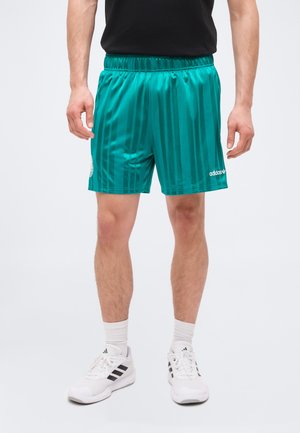 Mężczyzna w turkusowych sportowych szortach Adidas w paski, białych butach sportowych Adidas i białych skarpetkach, stojący na jednolitym tle.